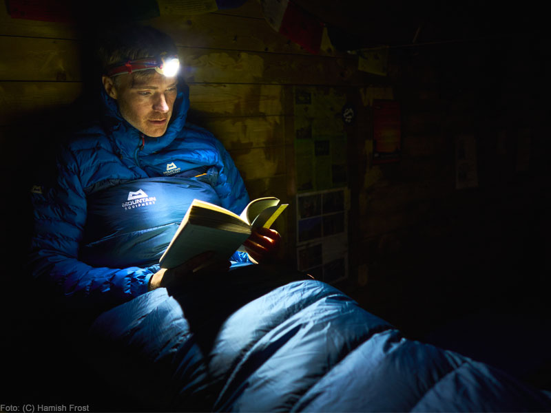 Mann mit Stirnlampe liest ein Buch in einem Schlafsack von Mountain Equipment.